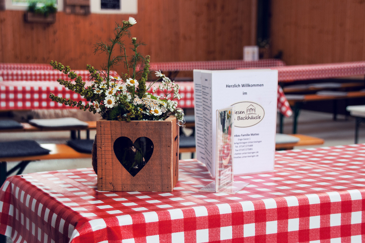 Ein Tisch mit rotem Karomuster ist dekoriert mit einem Holzkasten, der Blumen enthält, und einem Willkommenszettel für das "Bachhäusl Familie Mattes".