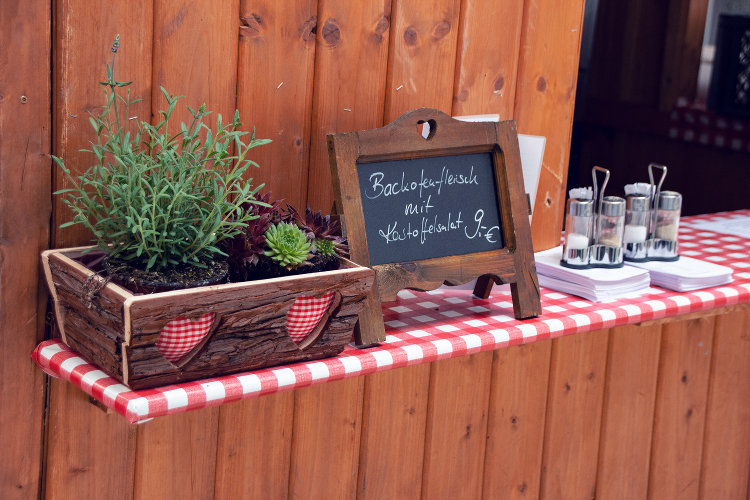 Ein rustikaler Tisch mit Holzbox, Pflanzen, Tafel mit der Aufschrift "Backofenfleisch mit Kartoffelsalat 9€" und Gewürzgläsern auf rotem-weiß kariertem Tischlaken.